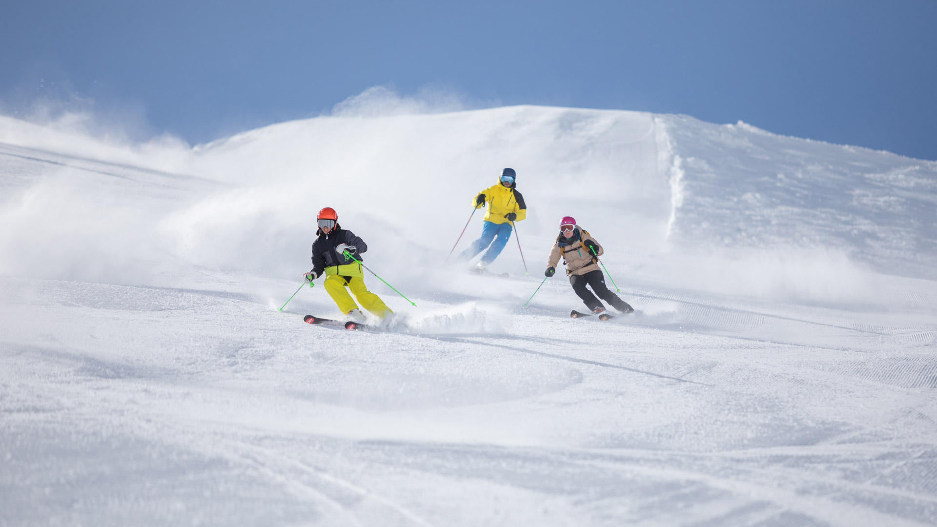 Drei Skifahrer gleiten schwungvoll über die perfekt präparierte Piste im Sonnenschein mit Pulverschnee.